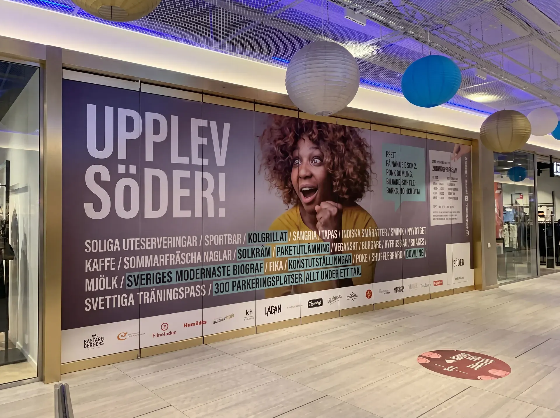 Stor reklamvägg med texten "UPPLEV SÖDER" och kvinna med lockigt hår i köpcentrum.